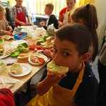 Chłopiec jje kanapkę, a z tyłu dzieci przy stole z warzywami.jpg.jpg